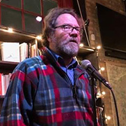 Errol McLendon with a graying beard wearing a plaid jacket blue button down shirt and glasses performing with a microphone in front of him and bookshelves behind him