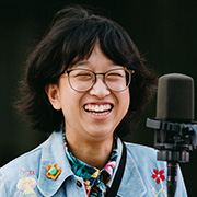 Jia Lian Yang smiling wearing dark colored rimmed glasses along with a light denium jacket with small flowers and performing with a microphone in front of them