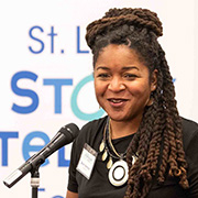 Shani Womack at a microphone performing at the St. Louis Storytelling Festival in 2023 wearing a black blouse and a large circle pendant necklace