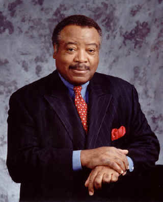 Julius K Hunter smiling wearing a light blue dress shirt with a red tie and dark colored suit with his right hand resting on top his left wrist