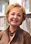 Joan Barry smiling with bookshelves behind her wearing a brown dress with a brown necklace