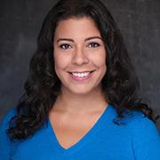 Jasmin Cardenas smiling and wearing a bright blue blouse with her long dark hair just below her shoulder against a gray background