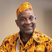 Kenya Ajanaku smiling while wearing a gold maroon and brown African dashiki and matching kente kufi hat