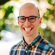 Adam Booth smiling wearing dark blue rimmed glasses, a multi-colored plaid shirt with trees and sunlight blurred in the background