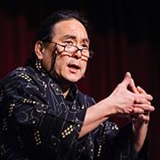 Alton Chung wearing glasses and Native American apparel performing on stage with hands folded out in front shoulder high