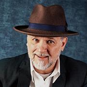 Simon Brooks with a graying beard wearing a white button down shirt, black sport coat and brown hat