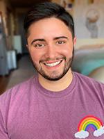 Aiden Thomas sporting a beard smiling and wearing a light colored maroon tshirt with a small rainbow and clouds on the left side of the chest