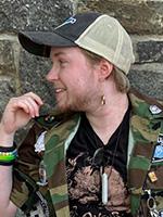 Andrew Joseph White with his head turned to the right and right hand at shoulder level sporting a black and white trucker hat and a green camouflaged jacket over a black printed t-shirt 