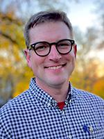 John Hendrix smiling and wearing dark rimmed glasses and a blue and white check button down shirt