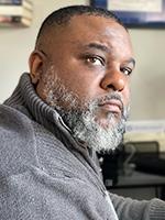 Lamar Giles looking up and to the right over his shoulder with a graying beard and sporting a gray high collared sweater