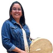 Storyteller Maria Hussman smiling and wearing glasses, a blue denium jacket, white tshirt while holding an Indian instrument