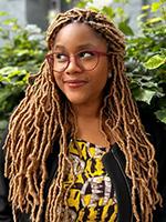 Namina Forna looking to the right with her long bleached braided hair down wearing glasses and yellow and black print top