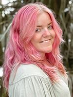 Samantha Markum with pink hair smiling and looking over right shoulder while wearing a white long sleeve top