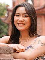 Trinity Nguyen standing against a red masonry wall with her arms folded and resting on the top and her long brown hair down